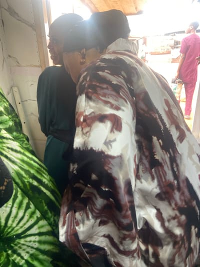 The image, apparently taken inside a building in Okengwe-Obehira, Nigeria, features a woman wearing glasses, a black cap, and a green top. She is positioned near a window, which is illuminating her face. Part of a patterned fabric drapes over her, obscuring much of her body. In the background, another person can be seen wearing pink clothing. The environment appears to be a simple interior with a white wall.