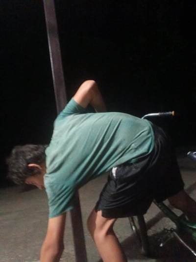 A person is bending over, touching a metal pole that appears to be a support structure. They are wearing a teal t-shirt and black shorts. The person has dark hair and is facing away from the camera. Their left arm is bent upwards, and their right arm is extended downwards towards the ground. A bicycle is visible in the background, with its handlebars illuminated. The scene is dimly lit, suggesting it is nighttime. The ground is a paved surface, possibly concrete or asphalt. The overall impression is of someone engaged in an activity outdoors at night, possibly adjusting or interacting with the bicycle or the pole. The location is specified as Leontyevskoye, Russia.