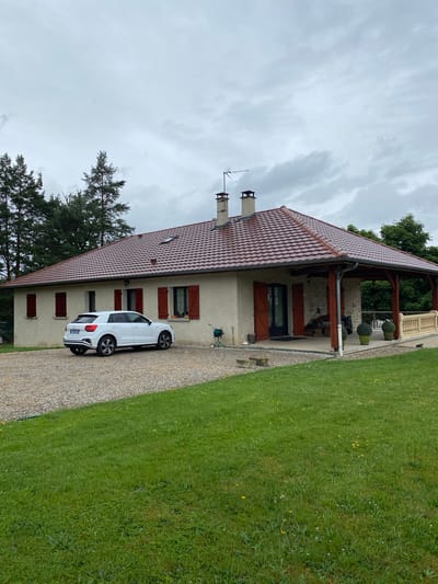 The image depicts a static outdoor scene featuring a two-story house, a parked car, and surrounding landscape under an overcast sky in Courpière, France. The house features light-colored stucco walls and a prominent reddish-brown tiled roof with two chimneys and a television antenna. Several windows are adorned with dark red wooden shutters.
A covered porch or veranda extends from the side of the house, supported by dark timber beams. It includes a balustrade and two potted topiary plants. A white SUV, identifiable as an Audi Q2, is parked on a gravel driveway alongside the house. The vehicle's rear license plate is visible, showing "FR" and "0787 JL".
The driveway borders a lush green lawn that appears slightly unkempt, scattered with yellow and white wildflowers. A large evergreen tree stands to the left of the house, and another deciduous tree is visible in the background. The sky is entirely covered by light gray, overcast clouds, indicating a cloudy daytime. A green garden hose reel is mounted on the exterior wall of the house near the porch. No human activity or specific events are taking place in the tranquil setting.