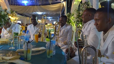 Five men in white suits sit around a table covered in a teal tablecloth. They are smiling and laughing, each holding a brass instrument, presumably trumpets or trombones. The table also holds various glasses, bottles of water, and clear plastic cups filled with drinks and straws. Floral arrangements and draped white fabric with twinkling lights adorn the background, suggesting a wedding or formal event.