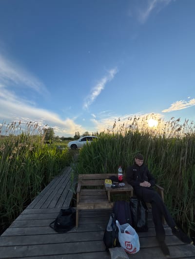 A man is lying on a wooden bench on a wooden platform, surrounded by tall reeds and grass, near a body of water, at dusk. The sun is low on the horizon. The sky is blue with scattered clouds. A white vehicle is parked nearby. On the bench next to the man are various items, including a bag, a water bottle, and other personal belongings. The photograph appears to have been taken in Khryashchevka, Russia.