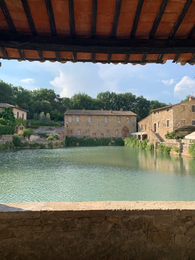 The image depicts a tranquil scene in Bagno Vignoni, Italy, likely during the daytime under a clear, partially cloudy sky. The perspective is from under a tiled roof structure, looking out towards a body of water. The water, a greenish hue, reflects the sky and the surrounding architecture.

On the far bank of the water, several historic buildings are visible, constructed from stone and featuring arched windows. These buildings are nestled into a lush, green hillside covered with trees and foliage. Terraced gardens with various plants and flowers add to the picturesque setting. Two of the buildings display flags, adding a touch of detail.

In the foreground, the underside of a terracotta tiled roof with exposed wooden beams is prominent, creating a sense of enclosure and framing the view. To the right, a rough stone wall transitions to a lighter, smoother surface, possibly the edge of a walkway or bridge, with the water lapping against it.

The overall impression is one of peacefulness and timeless beauty, characteristic of a historic Italian village. There is no discernible activity or event taking place, suggesting a moment of quiet observation. The lighting indicates it is daytime, with sunlight casting shadows and highlighting the textures of the stone and roof tiles. The scene evokes a feeling of serenity and immersion in a historical and natural environment.