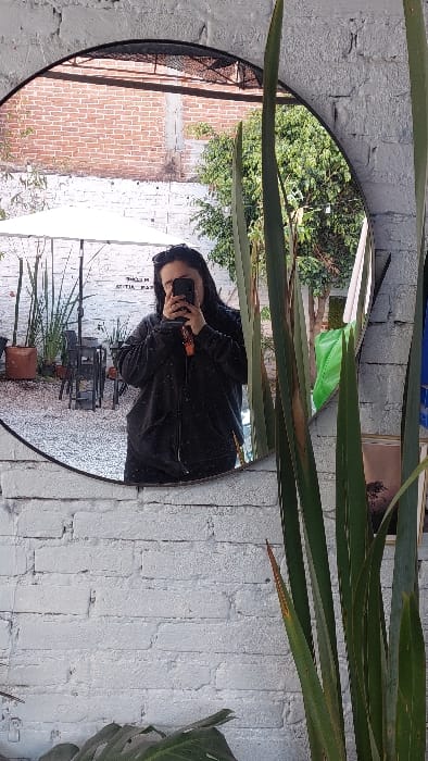 A circular, black-framed mirror, mounted on a white-painted, distressed brick wall, captures a reflection selfie within an outdoor commercial establishment in Tlaxcala City, Mexico. The reflection shows an individual, presumed female, with dark hair, wearing a dark zippered hoodie and sunglasses pushed onto her head. She holds a dark smartphone, featuring a visible orange charm, obscuring her face as she photographs her reflection. The environment reflected in the mirror includes an outdoor patio area with multiple dark metal tables and chairs positioned on a light-colored ground surface. A white umbrella or canopy provides shade. Various potted plants are arranged around the patio. In the upper background of the reflection, a red brick wall is visible, along with a dense green tree. A partially mirrored and inverted sign, possibly reading "WELCOME," is discernible. To the right of the mirror, outside the reflection, tall, sword-like green plants are prominent, and a large green leaf is visible at the bottom of the frame.