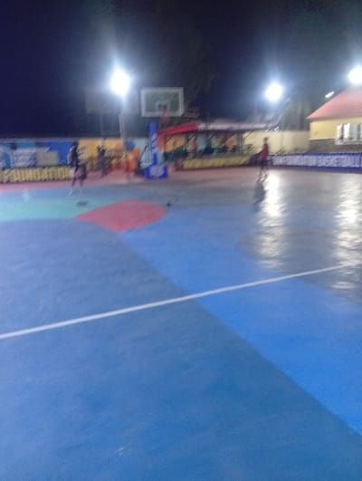 An outdoor basketball court under bright artificial lights at night. The court is painted in blue, red, and green sections, with a visible white line marking a boundary. In the background, there's a basketball hoop, banners, and blurry figures of people. A building with a red roof is visible in the distance. The overall scene is slightly blurry, suggesting a low-light photography setting.