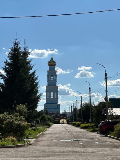 In the city of Mariinsky, Russia, a bright blue bell tower with a golden dome, topped by a cross, stands tall against a clear blue sky with scattered white clouds. The tower is flanked by a large, dark green coniferous tree on the left. A paved road leads towards the tower, with a row of streetlights on the right side of the road and lush greenery and bushes lining both sides. A red SUV is parked on the right side of the road, partially obscured by bushes. The road surface shows signs of wear and has several large puddles of water. In the background, beyond the streetlights, several houses with green roofs are visible.