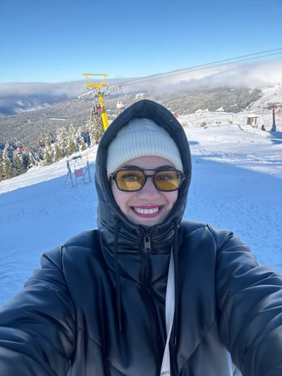 A young adult female is depicted in a close-up selfie taken at the Uludağ Ski Center, near Bursa, Turkey. She is smiling directly at the camera, revealing white teeth. She wears a black hooded puffer jacket, a cream-colored knitted beanie, and dark-framed sunglasses with yellow-tinted lenses. A white strap is visible over her right shoulder. The background consists of a vast snow-covered mountainous landscape under a clear blue sky with scattered clouds in the distance. A prominent yellow ski lift tower is visible, supporting cables on which multiple red ski lift chairs are suspended, some appearing occupied. Snow-covered coniferous trees populate the mountain slopes. Another smaller, reddish ski lift structure and a small wooden building are present in the far right background. The immediate ground in the scene is covered in snow.