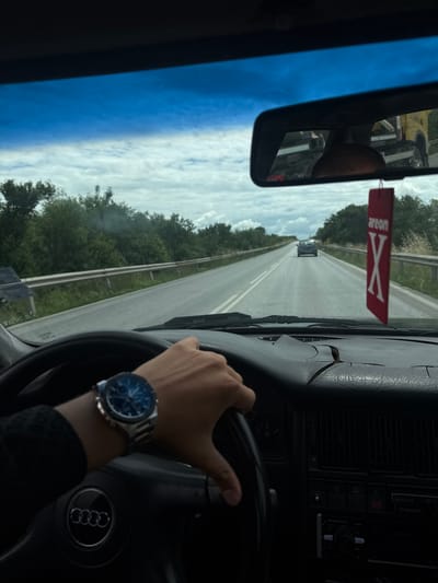 The image, taken from the driver's perspective inside a car, shows a cracked dashboard and a hand wearing a watch on the steering wheel. A red "Areon X" air freshener hangs from the rearview mirror, which reflects a yellow truck. The car is driving on a two-lane road lined with trees and a guardrail, with other vehicles visible in the distance. The sky is cloudy. The location is identified as the road entering Voyvodinovo, Bulgaria, near Plovdiv.