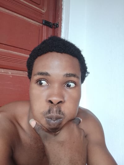 This media file features a close-up selfie of a young Black man. He is shirtless and resting his chin on his hand, with his fingers interlaced. His eyes are looking off to the side with a slightly surprised or curious expression. He has a light mustache and stubble. His hair is short and curly. The background shows a red, paneled door on the left and a plain white wall on the right. The lighting suggests it is daytime, possibly indoors or in a shaded outdoor area. The overall impression is casual and informal. There is no visible text in the image. The location context is Orodo, Nigeria.