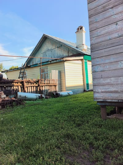 This is a medium-shot, eye-level, outdoor, full-color photograph of a rural dwelling in the village of Al'kino, Russia, on a clear day with blue sky and scattered clouds. The scene captures a section of a light yellow wooden house with a light blue weathered gable end, a silver metal roof, and a white brick chimney with visible openings at the top. A wooden ladder leans against the house. In front of the house is a dilapidated wooden fence with various metal objects, including what appear to be large cylindrical tanks, and piles of bricks and tires. The foreground is covered in lush green grass. To the right of the frame, a portion of a weathered wooden structure, possibly an outbuilding, stands elevated on concrete blocks.