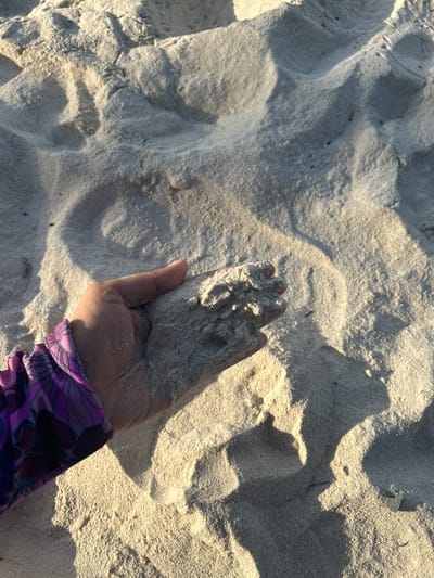 A close-up, overhead shot shows a person's hand holding a handful of light-colored sand. The hand is cupped, and the sand is slightly mounded within it. The person is wearing a purple rash guard with a floral pattern on the left arm, which is visible in the lower left quadrant of the image. The background consists of a vast expanse of rippled sand, likely a beach, with shadows cast by the uneven surface, indicating the presence of sunlight. The texture of the sand appears fine and soft. The overall lighting suggests it is either early morning or late afternoon. The scene is located on a sandy beach in A R A, Indonesia.