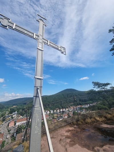 This image shows a tall metal cross positioned on a rocky outcrop overlooking the town of Dahn, Germany. The cross is inscribed with text, parts of which are legible and appear to read "Mitteln der K.-Kirche in L. errichtet 1872" which translates to "Erected by the middle of the K.-Church in L. 1872". In the background, a dense green forest covers a rolling hill, and the town of Dahn is visible below, featuring numerous houses with red-tiled roofs, a road with parked cars, and a small church spire. The sky above is a clear blue, dotted with scattered white clouds. A chain-link fence runs along the edge of the outcrop, separating the cross from the town and landscape below. The foreground is characterized by rough, reddish-brown rock formations and dry grass, with some puddles of water reflecting the sky.