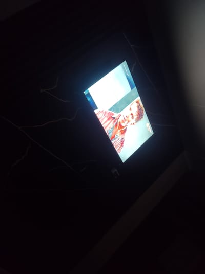 A medium shot captures a person's face on a tilted digital screen against a dark, possibly marble-patterned background. The person, appearing to be male, has light brown hair, fair skin, and is looking slightly to the left. He is wearing a striped red and white shirt. The screen displays what looks like a television show or movie, with a logo in the lower right corner reading "TVOO". The overall environment is dark, suggesting a nighttime or dimly lit indoor setting. The camera is positioned at an angle, and the screen itself is not perfectly flat against the surface behind it. There are no other visible people or significant objects in the immediate vicinity that would indicate a specific location within Қосшағыл ауылдық округі, Kazakhstan.