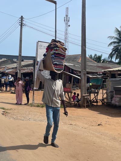 The image shows a man in Ikoga, Nigeria, balancing a large bundle of clothing on his head while walking on a dirt road. He is wearing a gray sweatshirt and blue jeans. In the background are buildings with corrugated metal roofs, utility poles and power lines, a cell phone tower, and other people. One sign reads "Power Char." Two children are sitting on the corner of a wooden structure. A woman in a pink outfit walks in the opposite direction.