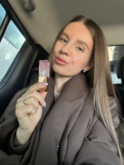 An adult female is depicted in a close-up, chest-up shot, seated inside a vehicle in the city of Zavidovo, Russia. She possesses long, straight light-brown hair and is looking directly at the camera with a subtle smile, her lips appearing glossy. She wears a dark brown, possibly padded, jacket over a lighter garment. Her left hand is raised, holding a transparent, rectangular bottle labeled "OPAL LIP OIL" with a gold-colored base and cap. Her left ring finger displays a two-banded ring with a clear stone. Her fingernails are manicured with light lavender polish featuring white stripe patterns. A small, heart-shaped stud earring with clear stones is visible in her right ear. The interior of the vehicle shows dark upholstery, and an out-of-focus view through a side window suggests an overcast or wet exterior environment.