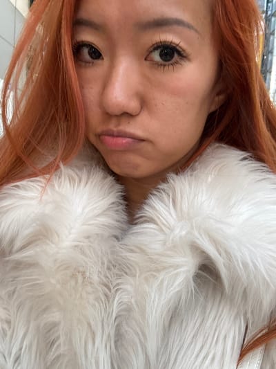 A woman with orange-red hair poses close to the camera. She has dark eyes, fair skin, and full lips. She appears to be wearing heavy mascara. Her head is tilted, and she rests against a fluffy, white faux fur coat or wrap. The background is indistinct and slightly blurred, but a glimpse of a white tiled floor suggests an indoor setting. The lighting is soft, highlighting the textures of the hair and fur.