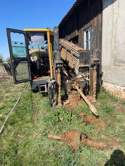 A compact drilling rig is positioned in a grassy area next to a building with a wooden facade and a brick foundation, possibly in a rural or semi-rural setting. The building features a window with a white frame. The drilling rig is yellow and black, with an open cabin and an extended drilling arm. A section of the drilling pipe lies on the ground, covered in mud. The immediate environment consists of grass and disturbed earth. The sky is clear and blue. The location is Поедуги, Russia.