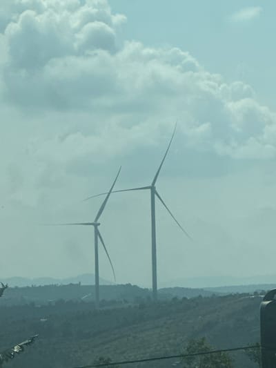 The image captures a landscape in Mui Ne, Vietnam, dominated by two prominent wind turbines silhouetted against a cloudy sky. The turbines, designed for renewable energy generation, stand tall with their blades extended. The foreground features a hilly terrain covered in vegetation, hinting at the natural beauty of the coastal region. The overall atmosphere is serene, with the soft, diffused light suggesting either early morning or late afternoon. The presence of wind turbines indicates a location in an area known for its wind power potential, consistent with Mui Ne's reputation as a popular tourist destination and windsurfing hub.