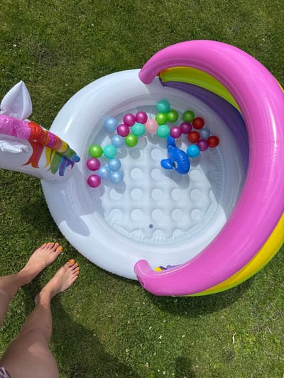 An aerial view shows an inflatable unicorn-themed kiddie pool filled with water and colorful plastic balls, set on a green, grassy lawn. A blue inflatable dolphin floats in the pool among the balls. To the left of the pool, two bare feet with bright orange nail polish are visible. The scene appears to be outdoors on a sunny day. The location is identified as Astrashitskagaradokski rural council, Belarus.
