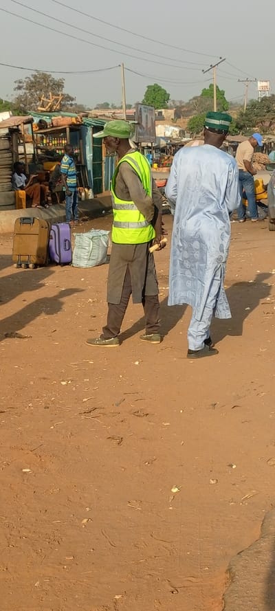 Two men stand on a brown dirt ground. One wears a bright yellow vest and a green hat, holding something in his hand. The other wears a light blue robe with a dark-striped green and blue hat. Suitcases and people sit near a roadside shop in the background. Power lines stretch across the sky above the scene.