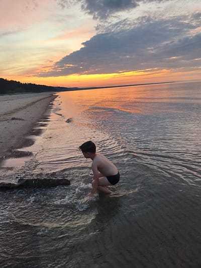 A young boy in black swim trunks is crouching at the edge of the Baltic Sea on a sandy beach in Apšuciems, Latvia. The sun is setting, casting a warm orange glow across the sky and reflecting on the calm water. The sky is filled with dramatic clouds, with a large, dark cloud formation extending from the right side of the frame. To the left, a dark line of trees borders the sandy shore. A piece of driftwood lies partially submerged in the shallow water near the boy. The water is clear enough to see the sandy bottom.