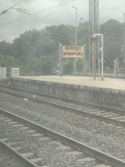 The image shows a train station platform with railway tracks in the foreground. A sign board on the platform reads "BANAPURA" in English and "बानापुरा" in Hindi, indicating the location is Banapura, a town in the Seoni Malwa tehsil of the Narmadapuram district in the Indian state of Madhya Pradesh. The sky is overcast, and lush green trees are visible in the background. Two individuals are seated on the platform in the middle ground, one appearing to be talking to the other. Several electrical poles and wires are also visible. The platform appears to be made of concrete, and the railway tracks are laid on ballast.