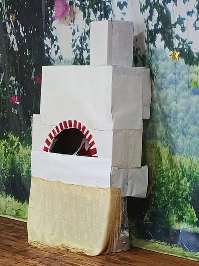 This media file shows a handmade model of a traditional Russian stove, known as a "pech". The stove is constructed from white cardboard boxes of various sizes stacked and glued together to mimic the layered structure of a real pech. The opening of the stove is decorated with red and white stripes forming an arch. The base of the stove is covered with a textured, light yellow material that resembles straw or dried grass, with frayed edges hanging down.

The stove is placed in front of a backdrop that appears to be a fabric print or a mural depicting a forest scene with trees, foliage, and hints of blue sky. There are a few small, colorful elements in the backdrop, including pink flowers on the left and a shiny gold star hanging on the right, suggesting a festive or decorative context, possibly for a play, a diorama, or a craft project. The lighting is consistent, and there are no strong shadows, indicating it's likely an indoor setting with artificial light or overcast daylight.

The overall impression is of a charming, handcrafted model representing a significant element of traditional Russian rural life. There is no visible text in the media file. The absence of people and the static nature of the object suggest it is a still life composition or a prop.