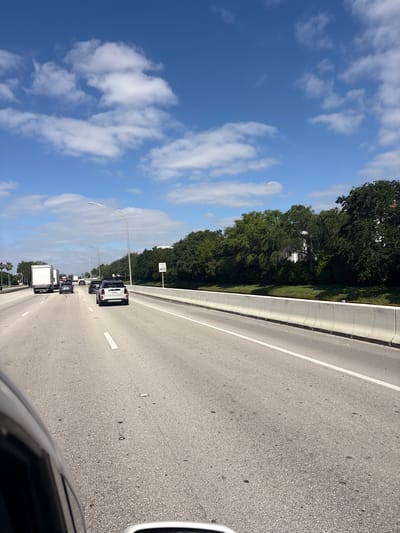 A daytime photograph captures a view from a vehicle on a multi-lane highway, looking forward into moderate traffic. The sky, a vibrant blue with scattered white cumulus clouds, fills the upper left quadrant of the image. Along the right side of the highway, a sturdy concrete barrier separates the road from a dense stretch of lush green trees.

The asphalt road features clear white lane markings. Several vehicles are visible ahead, suggesting a busy flow of traffic. Prominently in the rightmost lane, a white SUV with a dark roof leads a line of cars, followed by various sedans and a white box truck further down the road. A dark-colored vehicle, likely the one from which the photo was taken, is partially visible in the bottom right corner, showing its hood and side mirror.

Tall streetlights with horizontal arms are spaced along the highway. A rectangular white speed limit sign, mounted on one of these poles on the right, clearly displays "SPEED LIMIT 40". The bright sunlight indicates a clear day. The scene depicts a typical highway environment in an urban or suburban area like Doral, United States of America, with traffic moving under sunny conditions.