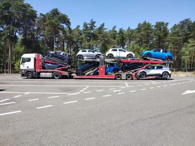 The image depicts a car carrier truck loaded with multiple vehicles parked on the side of a road with a forest in the background. The truck is white and red and is carrying eight cars in various colors including black, silver, white, and blue. The cars are stacked on two levels of the carrier. The road is asphalt and has white lane markings. The environment is a bright, sunny day with a clear blue sky. The location is identified as Theillay, France.