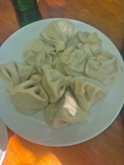 A top-down shot shows a white plate filled with approximately a dozen Khinkali (Georgian dumplings) resting on a wooden surface, possibly a table. A dark green bottle is partially visible on the lower left. The light suggests an outdoor or well-lit environment. No people are in the view. No location information is available.