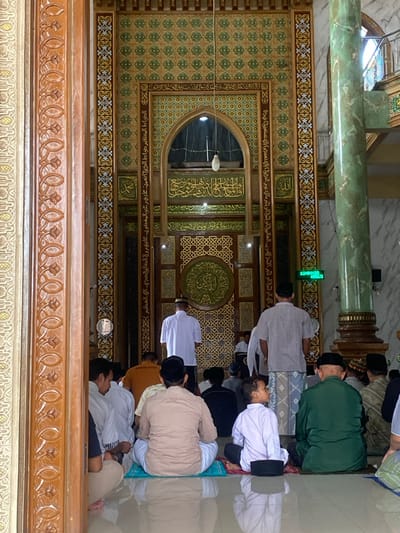 This image depicts the interior of a mosque located in the city of Jepara, Indonesia.
Approximately 15 to 20 male individuals, ranging from young boys to older men, are present. Most are seated on a highly polished, light-colored floor that exhibits strong reflections. Several individuals are seated on small, rectangular prayer mats in light blue, red, and green. Their attire includes long-sleeved shirts (white, beige, green, orange, striped), patterned sarongs, and black peci caps. Two adult men are standing with their backs to the viewer, facing the front wall: one in a white shirt and sarong, and another in a striped shirt and patterned sarong. A young boy in a white shirt and black peci is seated in the foreground, looking to his left. The seated individuals generally face forward towards the ornate mihrab area.
The front of the mosque is characterized by elaborate architectural detailing. It features a decorative wall with rich brown wood, intricate gold and green geometric patterns, and extensive Arabic calligraphy in gold. A central arched recess near the top of this wall contains a single white pendant light fixture. To the far left, a carved wooden column displays a repetitive, organic circular motif. To the right, a prominent, fluted column made of a green marble-like material stands on a golden base. Behind this green column, a white tiled or marble wall is visible, along with a small, green digital clock displaying "12:00:41". A section of an upper-level balcony with a metal railing is partially visible in the upper right corner. The space is brightly lit, illuminating the reflective floor and detailed surfaces.