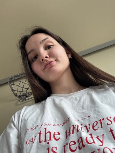 The image presents a close-up, low-angle selfie of a young adult female with long, dark brown hair and fair skin, looking directly at the camera with a neutral expression. She is wearing a white t-shirt with red lettering, part of which reads "just remember" in a cursive script, and "04/ the universe is ready to" below it. A small, gold-colored hoop earring with a clear stone is visible in her right earlobe. The background consists of a plain, light beige wall with a horizontal gray trim. To the left of the subject's head, a wall-mounted silver-colored metal mesh rack, secured by two screws, is visible. This interior environment suggests a public transportation vehicle, such as a train car. The image contains no specific visual markers to identify a precise location within the city of Barabinsk, Russia, beyond being an indoor setting consistent with travel in that geographical area.