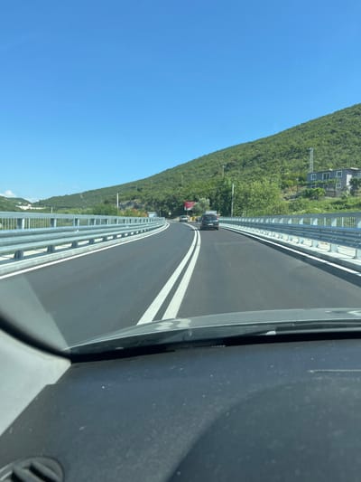 The image shows a view from inside a vehicle driving on a highway in Kruče - Kërruçi, Montenegro. The road is paved with asphalt and has white lane markings. There are guardrails on both sides of the road, made of metal. The highway is bordered by a lush green hillside on the right and a slightly less dense hillside on the left, with some scattered buildings visible on the right hillside. The sky is clear and blue. Two cars are visible on the road ahead, one in each lane. The interior of the vehicle, including the dashboard and windshield, is visible in the foreground.