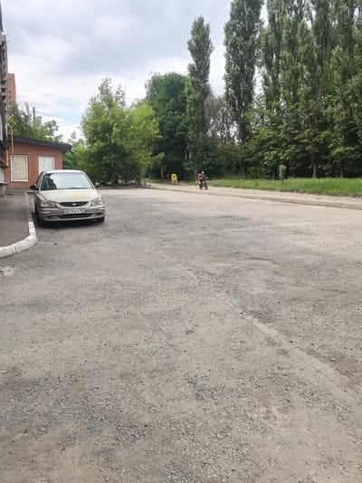 A light-colored sedan is parked on a gravel lot next to a brick building. The car, a Hyundai Accent, has a license plate that reads "M214AX 161". To the right of the car, a person on a motorcycle with a red backpack is moving away from the camera. In the background, a line of tall trees and shorter green foliage forms a border between the lot and a grassy area. The sky is overcast. The scene appears to be in an urban or semi-urban area, potentially a parking lot or side street, possibly located in Верхнетемерницкий, Russia, based on the license plate format.