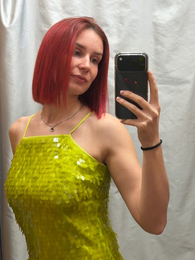 A woman with red hair is holding up a phone, taking a selfie. She is wearing a green sequined dress and a silver necklace. Her hair is a vibrant red, and she has fair skin. She has a black hair tie on her wrist. The background is a light gray or white material.
It is impossible to verify if this image was taken in Зюкайка, Russia, as there is no information to confirm the location.