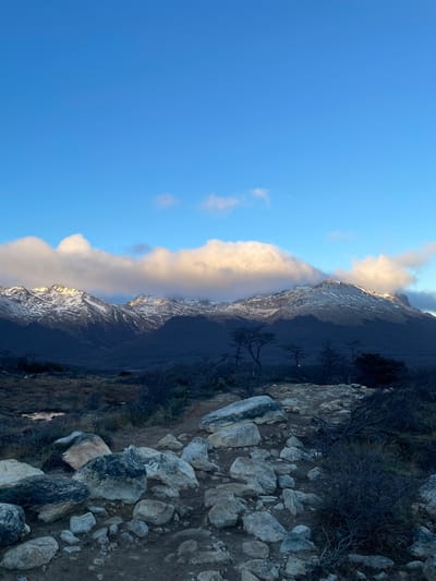 The image shows a mountain landscape in an unknown location in Argentina. The foreground depicts a rocky hillside with sparse vegetation. In the background, a tall, dark mountain range is visible, partially covered with snow and a long strip of clouds. The sky above is a clear blue.