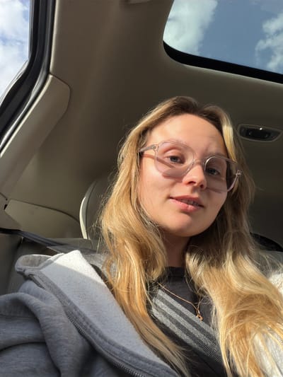 A selfie-style, close-up photograph depicts a light-skinned young woman with blonde hair and clear-framed glasses inside a vehicle. The vehicle's roof, including a sunroof showing a cloudy sky, is visible. The woman wears a dark shirt and a gray hoodie with a visible zipper. A thin, gold necklace is also visible on her chest. The interior of the car appears neutral in color. The specific location in Arava Tichona Regional Council, Israel, cannot be determined from the image.