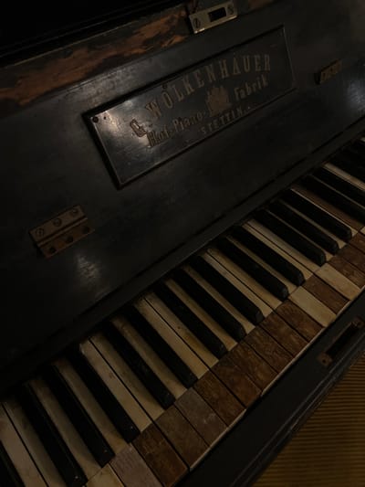 This is a close-up, high-angle photograph of an antique upright piano. The piano is dark, possibly black, and appears aged with wear marks visible on its surface, particularly near the top. A metal plaque is affixed to the upper portion of the piano, bearing the inscription "C. WÖLKENHAUER Hof-Piano-Fabrik Stettin". The keys of the piano are visible, with a mix of black and ivory (or aged white) keys. Many of the ivory keys show significant discoloration and damage, with some appearing to be missing or replaced with wooden pieces. The piano is situated on a floor covered with a striped rug, likely in a residential setting. There are no people or animals present in the image. The scene is dimly lit, creating a somber and nostalgic mood. The location of Beit Hanan, Israel, is irrelevant to the visual content of the image itself.