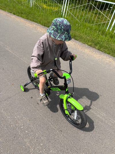 A young child wearing a bucket hat and a grey jacket rides a bright green bicycle with training wheels on an asphalt path. The child is seen from a high angle, looking down at the bicycle's handlebars. The path is bordered by a grassy area with a white metal fence and scattered yellow flowers. The scene appears to be outdoors in a rural or park-like setting.