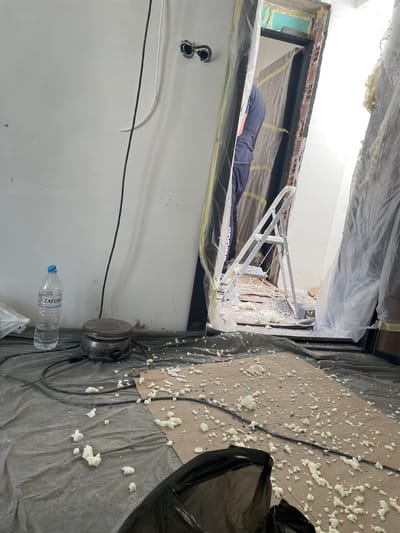 The image depicts an indoor construction or renovation scene, likely in Skala Potamias, Greece, based on the visible text. The floor and surrounding areas are covered with grey plastic sheeting, secured with yellow tape, and partially with cardboard, to protect against spills and debris. Numerous dried white and yellowish blobs of spray foam insulation are scattered across these protective covers.

In the center, a person wearing a dark blue top and pants is lying prone, reaching into a window frame or wall opening. Part of the person's clothing has "adidas" faintly visible. The opening reveals exposed red brickwork and some green insulation material, with fresh spray foam applied around the frame. A metallic, ladder-like structure is positioned inside the opening, near where the person is working.

On the floor, near the wall, an electric hot plate is visible next to a clear plastic water bottle with "ZAGORI" printed on its label, a Greek brand of bottled water. A black electrical cord runs from the hot plate across the protective sheeting. Another black trash bag is partially visible in the foreground, and white and black electrical wires run along the lower wall. The lighting suggests it is daytime.