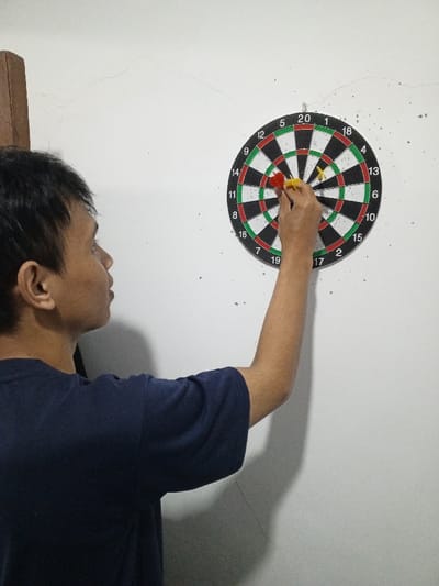 An adult male, facing right in profile, is depicted indoors in Al Haouafate, Morocco, interacting with a dartboard mounted on a white wall. He wears a dark blue short-sleeved t-shirt. His right arm is extended, and his hand grasps two darts embedded in the dartboard: one red-fletched dart in the outer segment of the '20' area, and one yellow-fletched dart in the single segment of the '1' area. A third yellow-fletched dart is visible stuck in the single segment of the '4' area. The dartboard is a standard numbered target, featuring black, white, red, and green sections. The white wall exhibits minor cracks and scattered dark specks, possibly impact marks. A dark brown vertical wooden structure is partially visible on the far left.