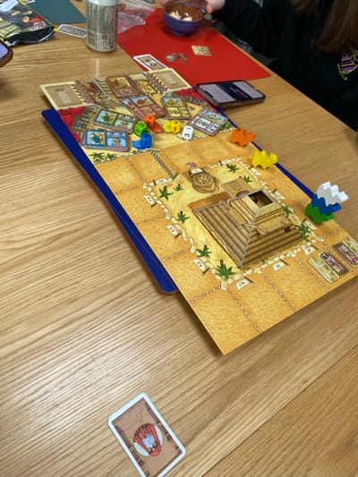 The image shows a game of the board game "The Mummy" being played on a wooden table. The game is in progress and players have placed dice, cards, and game pieces on the board. There are several dice, some of which are colored, and several cards, one of which is in the bottom right corner of the image. There are also plastic game pieces, and a yellow plastic pyramid, on the board. The board is designed to look like an Egyptian pyramid with pathways around the outside and a central chamber. The game is being played on a wooden table, which appears to be in a home setting. A person is sitting at the table, but only their arm can be seen in the image. Based on the coordinates, the image was likely taken in Keswick, a town in the Lake District in the United Kingdom.