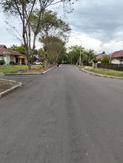 The image depicts a street in Palohgadeng, Indonesia, lined with trees and residential buildings. The road is paved and appears to be in good condition. Several houses with red tile roofs are visible on the right side of the street, separated from the road by a fence and vegetation. On the left side of the street, more trees and buildings are present, including what appears to be a white SUV parked in front of one of the houses. The sky is overcast with gray clouds. Two individuals, one in a pink outfit and the other in a white top and black pants, are walking away from the camera down the street on the right side. Another individual in a white top and dark pants is walking towards the camera on the left side of the street. A street light is visible on the right side of the road. The foreground is dominated by the asphalt of the street, with some debris and a curb visible. The overall impression is a quiet, residential street scene on a cloudy day.
