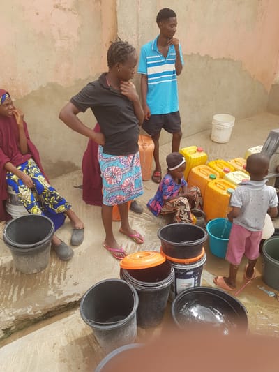 The image shows a group of individuals, including children and adults, positioned on a concrete or tiled surface in Dutsinma A, Nigeria. They are surrounded by yellow plastic jerrycans and several black buckets, some of which appear to contain water, indicated by the presence of reflections. One child is pouring water from a blue bucket. Some individuals are seated or lying down, while others are standing. The environment is slightly wet, suggesting a recent spillage. Clothes are colorful, and a white bucket is seen in the background near a wall.