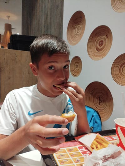 A young boy with short, dark hair is eating a chicken nugget. He is wearing a white Nike t-shirt with a blue logo. He is sitting at a table with a tray of food, including french fries and a drink. The background appears to be a restaurant with wooden wall decorations. The boy is the sole subject of the image and is focused on eating. The scene suggests a casual dining experience.