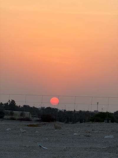 The image depicts a sunset over Umm Salal Muhammad, Qatar. A large, vibrant orange sun is partially obscured by a wire fence that spans the foreground. Beyond the fence, the landscape is arid and rocky, with scattered debris. A line of trees, including palm trees, forms a dark silhouette against the warm-toned sky. In the mid-ground, there are several low-lying buildings and structures, some appearing to be in disrepair. Further in the distance, what appear to be telecommunication towers and streetlights are visible. The sky is a gradient of soft orange and pink, characteristic of a sunset. No people are visible in the scene.