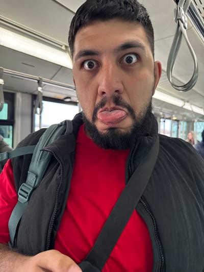 A man with dark hair and a beard is pulling a silly face with his tongue sticking out and eyes wide open. He is wearing a red shirt and a black jacket with a grey backpack slung over his shoulder. The background suggests he is inside a train or bus with rows of windows and overhead hand grips visible. The image was taken from a close-up, slightly tilted perspective. The location in the city of Емельяново (аэропорт), Russia, cannot be determined from the image alone.