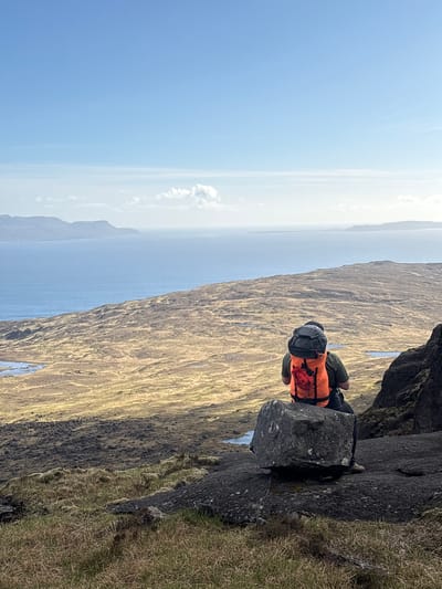 A hiker is resting on a rock ledge on the Isle of Skye, United Kingdom. The hiker wears an orange vest, green pants and carries a grey backpack. The hiker is positioned on a grey rock face. To the left, there is a view of water and distant land under a blue sky. To the right, the rock face is covered in dry grass.