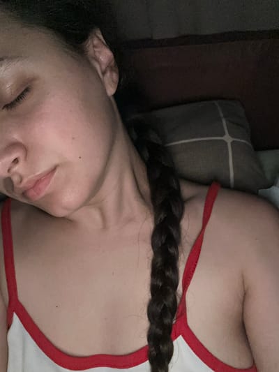 A close-up, dimly lit photograph shows a person with their eyes closed, appearing to be asleep. Their head is tilted, resting on a pillow with a patterned cover. The person has dark hair braided and falling over their shoulder. They are wearing a white tank top with red trim. The scene is indoor and appears to be a bedroom. There is no identifiable information to suggest a location in Postoyata, Ukraine.