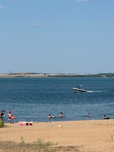 The image depicts a sunny day at a beach or lakefront, likely in Антонов ауылдық округі, Kazakhstan, given the provided context. The scene is divided into a sandy beach area on the right and a large body of blue water on the left. A clear blue sky with some faint clouds suggests fair weather.
Several people are visible in and around the water. On the sandy beach, individuals are lounging and sunbathing, some on towels. In the water, a few people are swimming or wading. A small white boat is actively moving across the water, leaving a wake, indicating recreational activity.
The landscape in the background shows a treeline along the shore, with what appear to be power lines and pylons further inland, suggesting a developed or semi-developed area. The time of day appears to be afternoon, given the bright sunlight casting shadows. The overall mood conveyed is one of relaxation and enjoyment of a summer day. No visible text is present in the image.