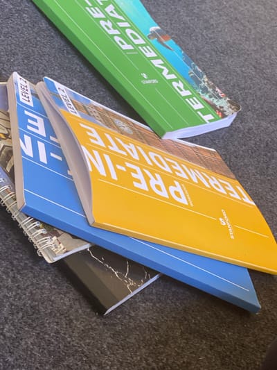The image shows a stack of books on a dark, textured surface, likely carpet. The top book is green with white text that reads "PRE-INTERMEDIATE TERM." Below it is a yellow book with white text that reads "PRE-INTERMEDIATE WORKBOOK." Underneath the yellow book is a blue book with white text that appears to be "LEVEL 2 E-IN". The corner of another book, possibly a notebook with a marble pattern, is visible at the bottom. The scene is indoors. There are no people or animals present. The lighting suggests an indoor setting. The image does not provide any information about the city of Arbagish, Uzbekistan, or its specific location within that city.